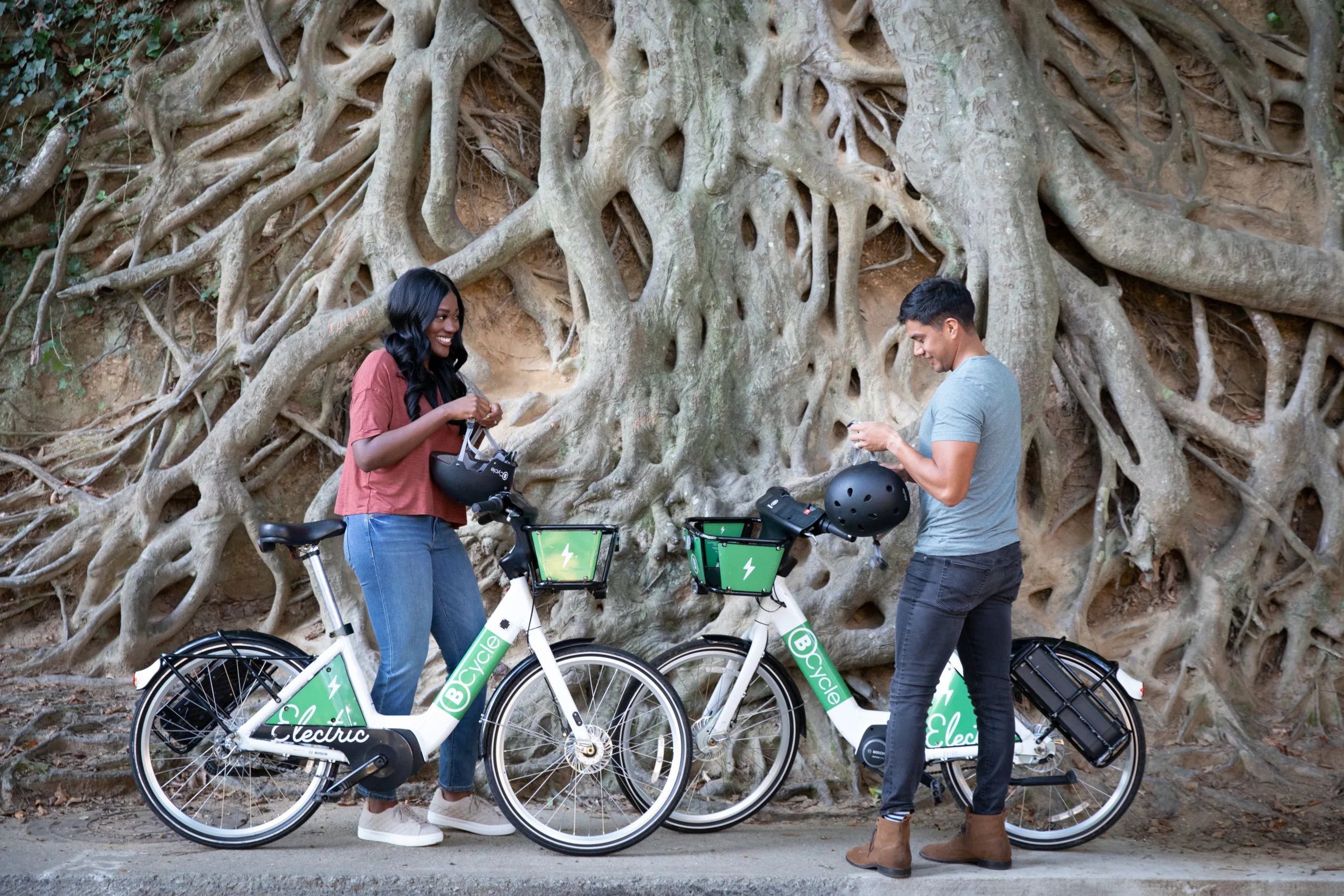 Woman and man standing in front of large tree and it's roots on bike share bicycles