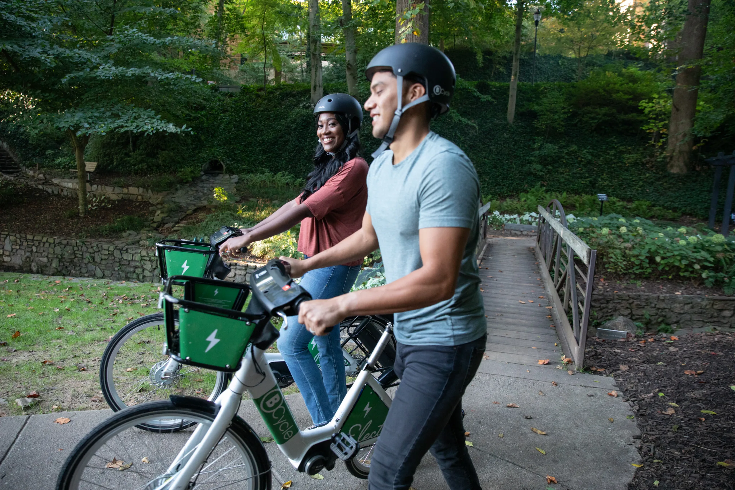 Black woman smiling at camera walking her bicycle on the Swamp Rabbit Trail with a latino man